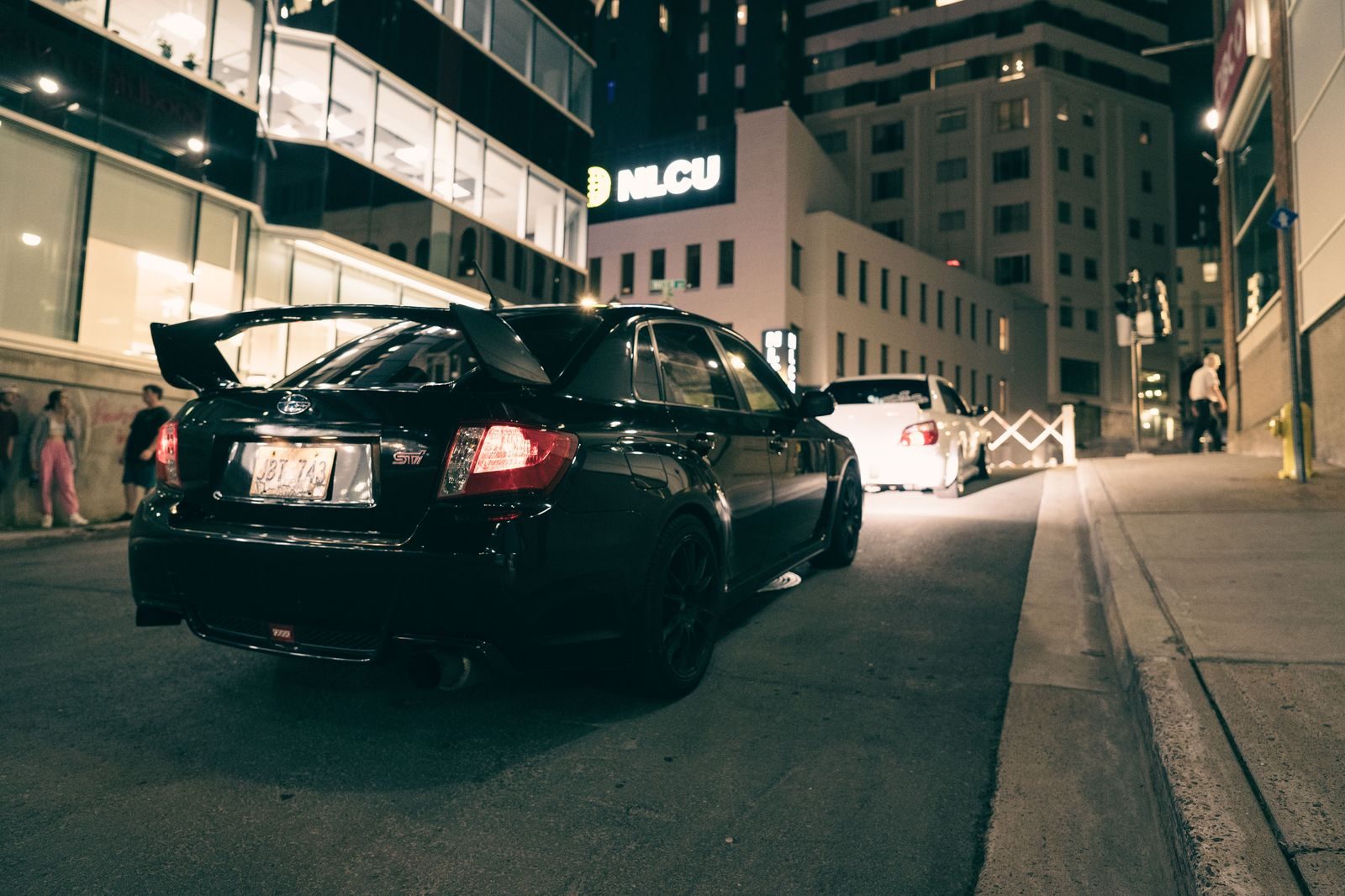 A black sedan waiting at a rainy kerb at night outside a cinema